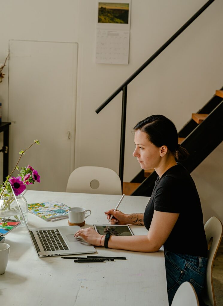 Woman in Black Crew Neck T-shirt Sitting by the Table While Using Laptop